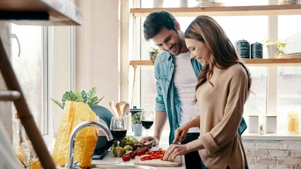 Young couple smiles as woman chops vegetables and man leans in, watching.