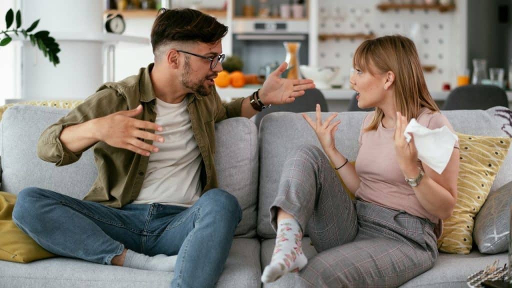 A man and woman sit on a sofa, gesturing expressively during a heated discussion.