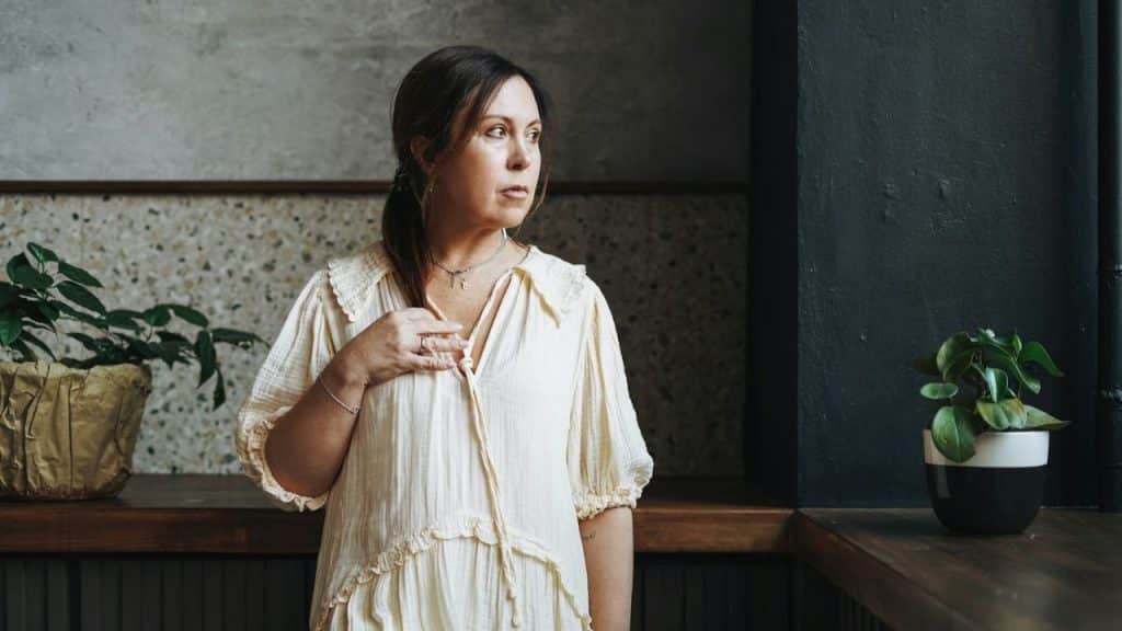 Middle-aged woman in beige dress stands looking out, next to dark framed window.