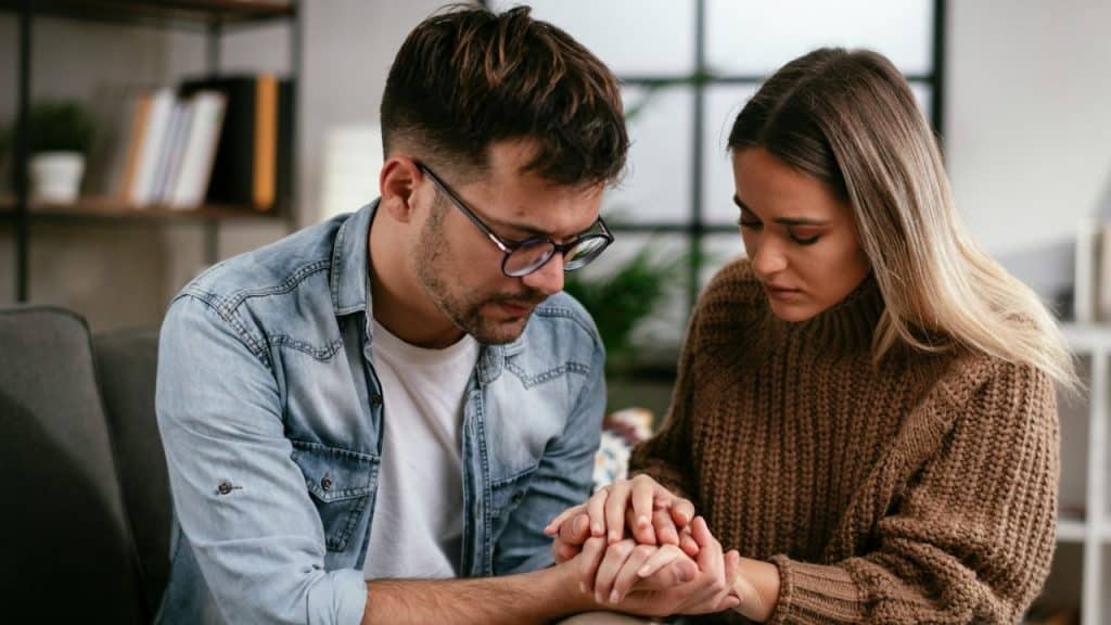 Young couple sits on couch, man holds woman's hands, both looking down sadly.