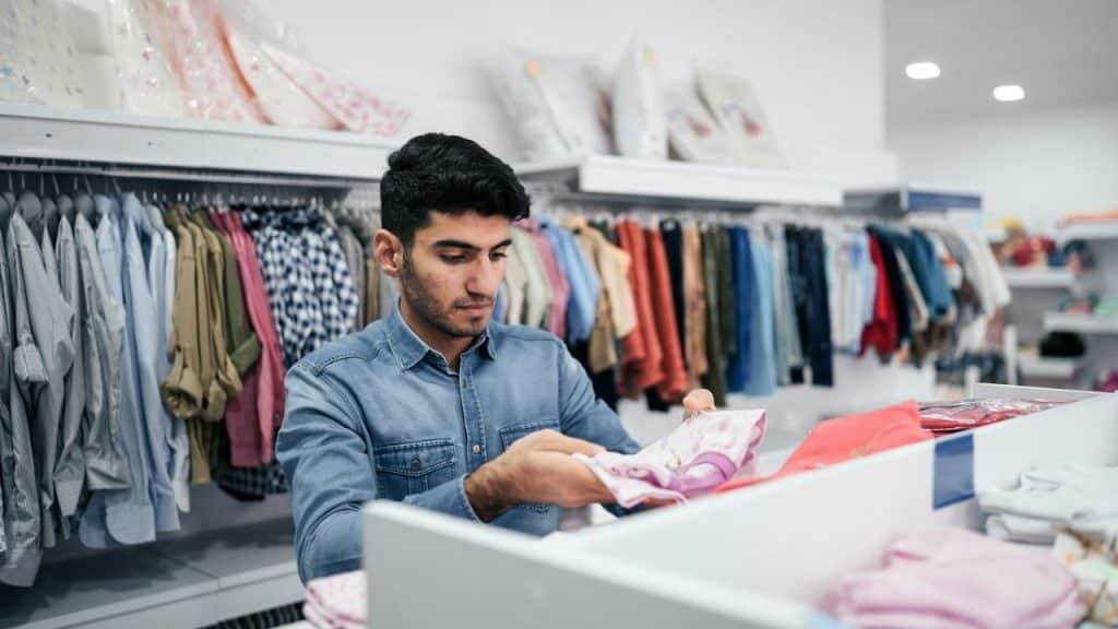 Man in a denim shirt examining clothes in a brightly lit retail store.