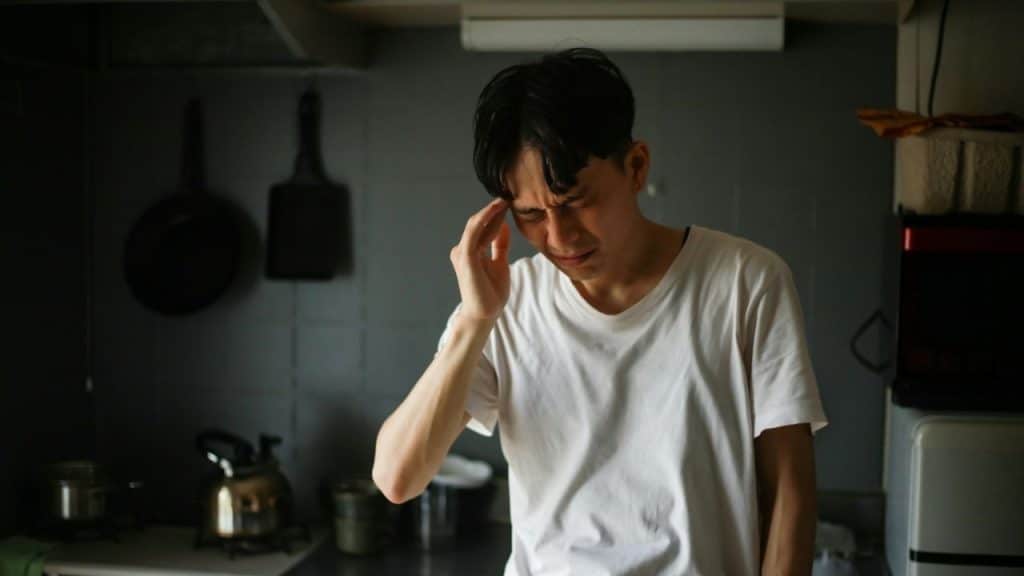 Man in a white t-shirt holding his head in a dimly lit kitchen with cooking utensils.