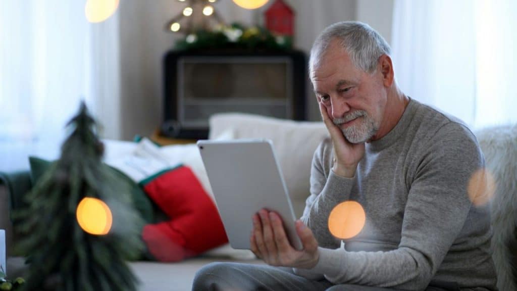 Older man in gray shirt looking at a tablet with a sad expression in a decorated room.