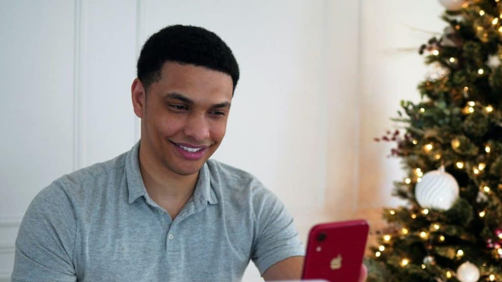Man in a gray shirt smiling while looking at a red smartphone near a Christmas tree.