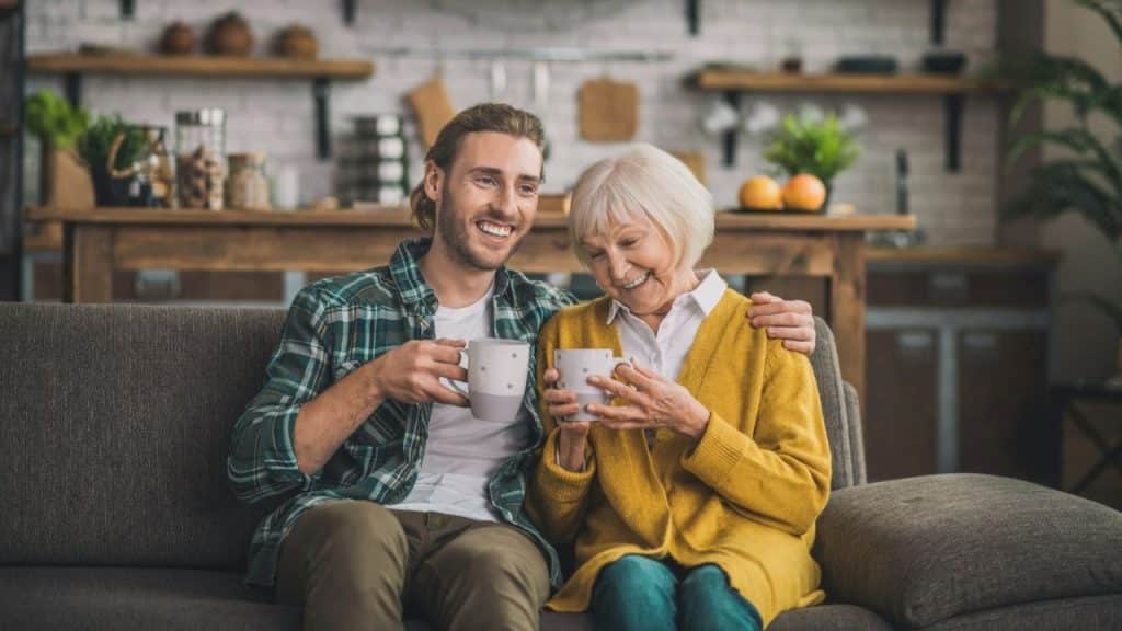 Young man and older woman sitting on a couch, holding mugs and smiling.