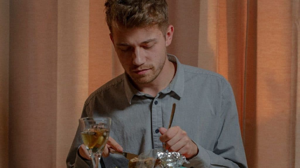 Man in a gray shirt cutting food with a fork and knife, with a glass of wine.