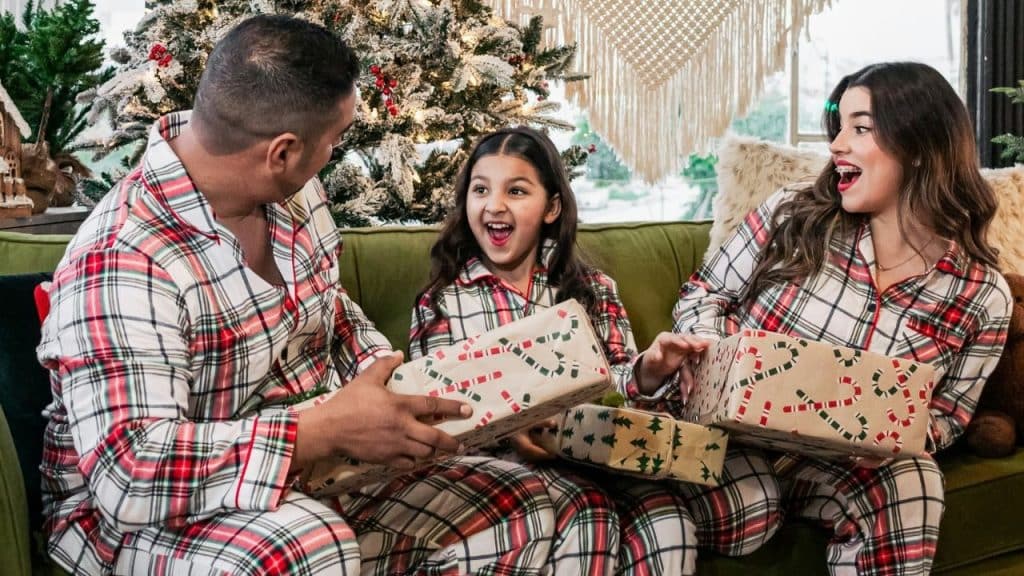 Three people in plaid pajamas happily opening gifts on a sofa near a Christmas tree.