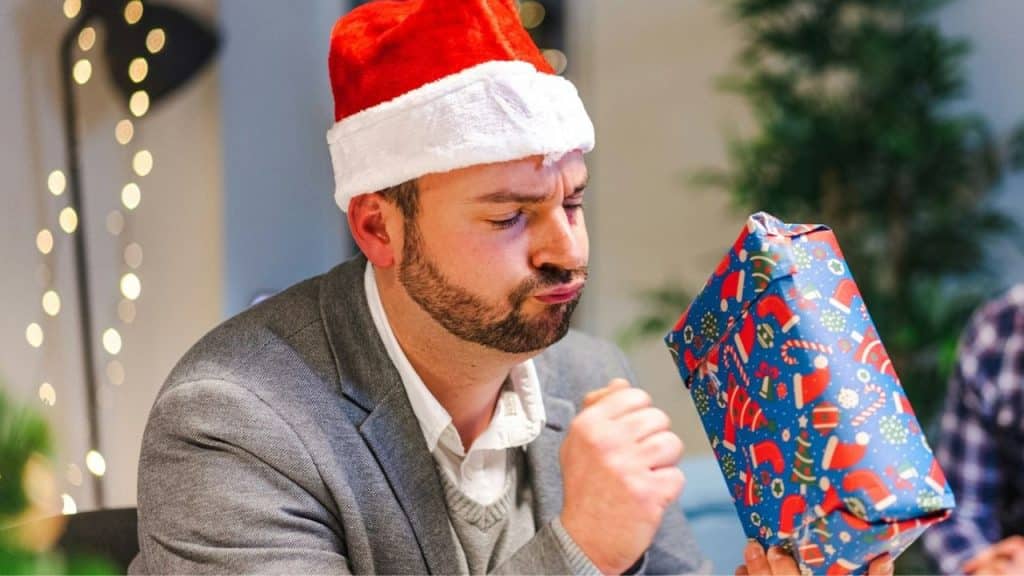 Man in Santa hat looking curiously at a blue, patterned wrapped gift he is holding.