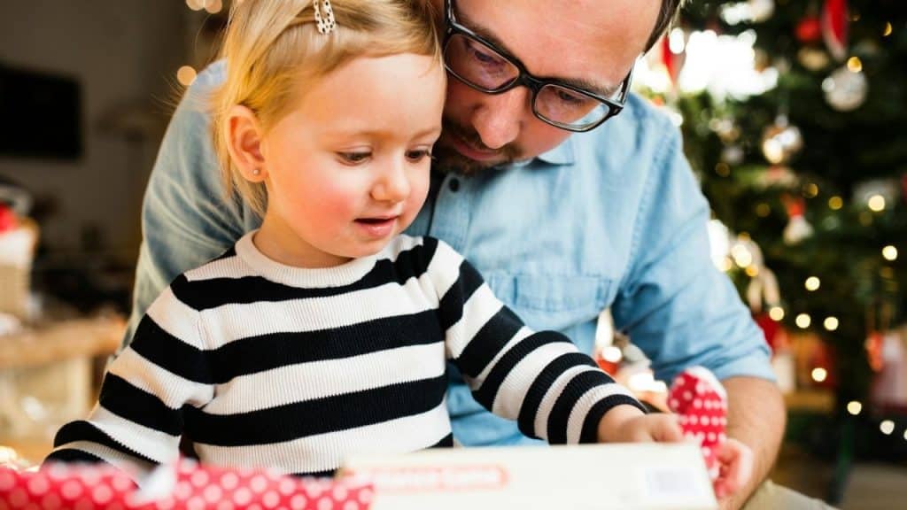 Man with glasses helping child in striped shirt open a red polka-dot gift.