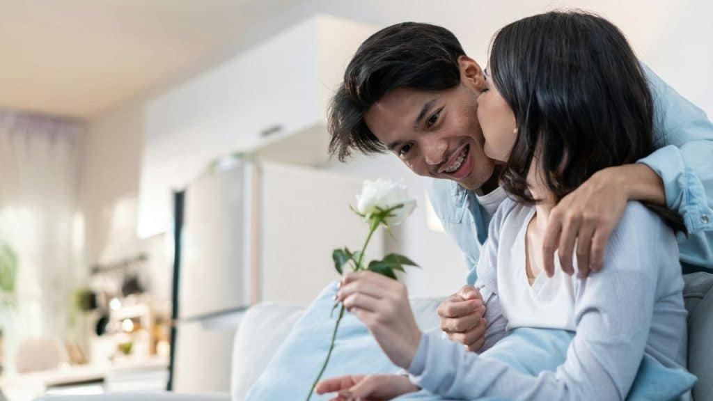 Young man hugging woman from behind while giving her a white rose indoors.