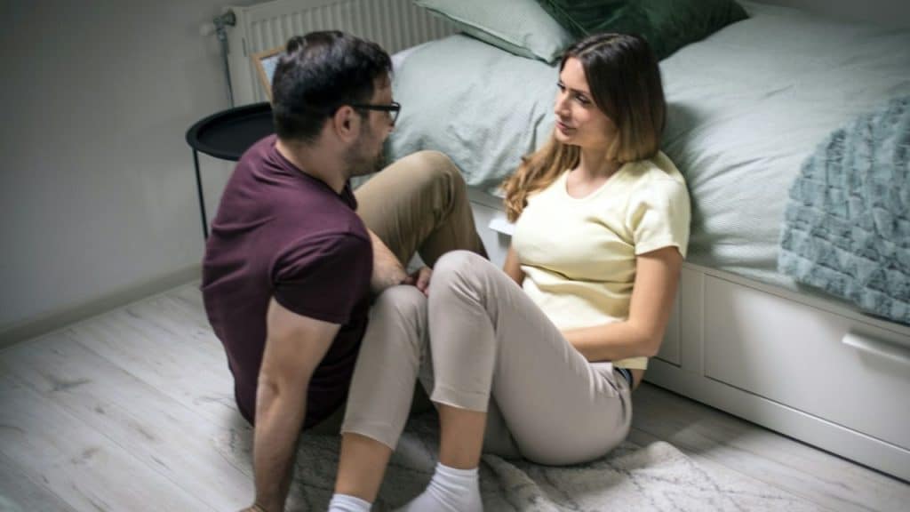 Couple sitting on the floor leaning against a bed, talking and looking at each other.