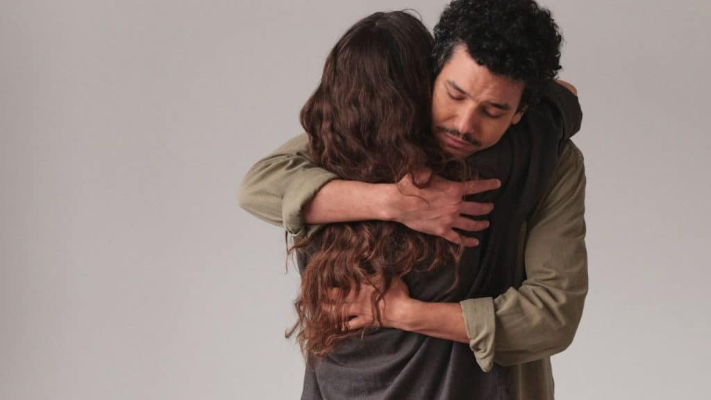 Man with a mustache hugging a woman from behind against a plain gray background.