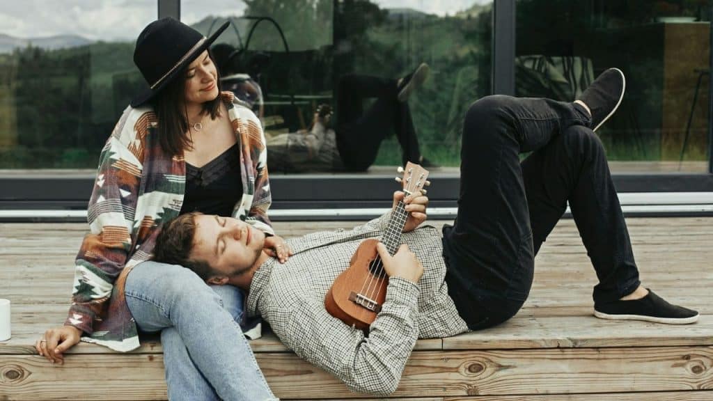 A woman sitting with a man lying on her lap playing a ukulele outdoors.