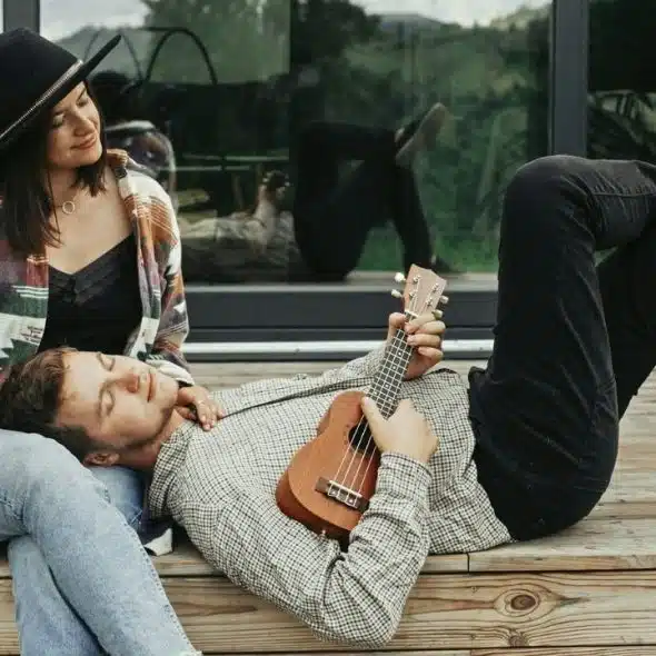 A woman sitting with a man lying on her lap playing a ukulele outdoors.