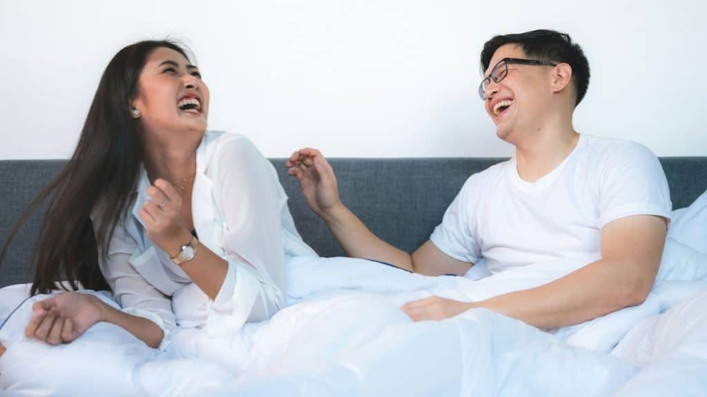 Man and woman laughing happily in bed with white sheets and a gray headboard..
