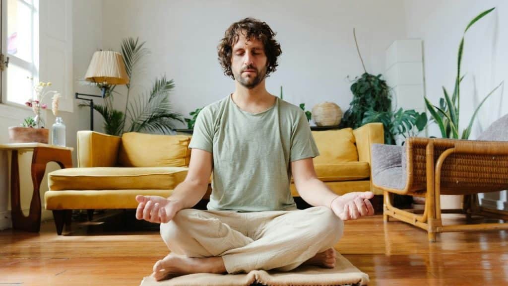Man sitting in cross-legged meditation pose on a mat in a bright living room.