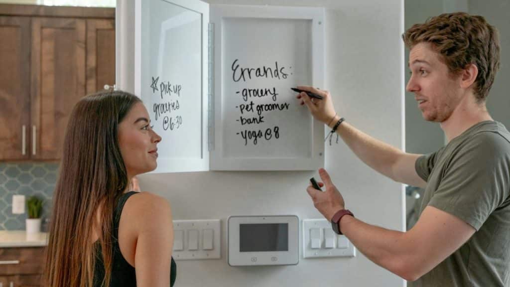 Man writing a list of errands on a dry-erase board while a woman looks on.