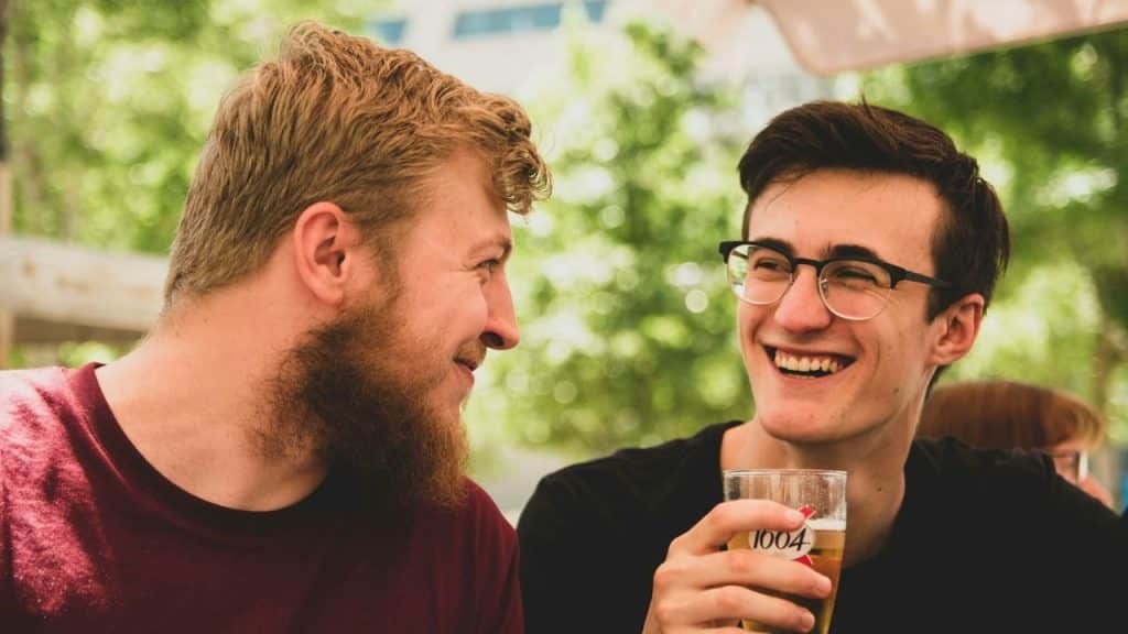 Two men sitting outside, smiling and talking, with one man holding a glass of beer.