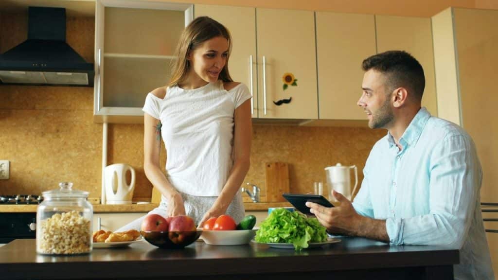 Smiling couple talking in a kitchen, with a tablet and fresh produce on the counter.