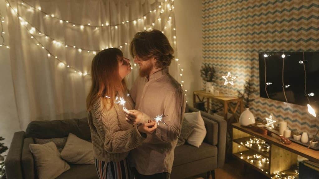 A couple holding sparklers and standing close together in a warmly lit, festive room.
