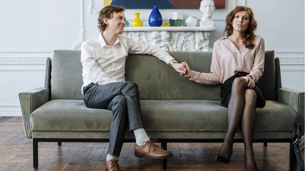 A man and a woman sit on a grey sofa in a brightly lit room, holding hands and looking at the camera.
