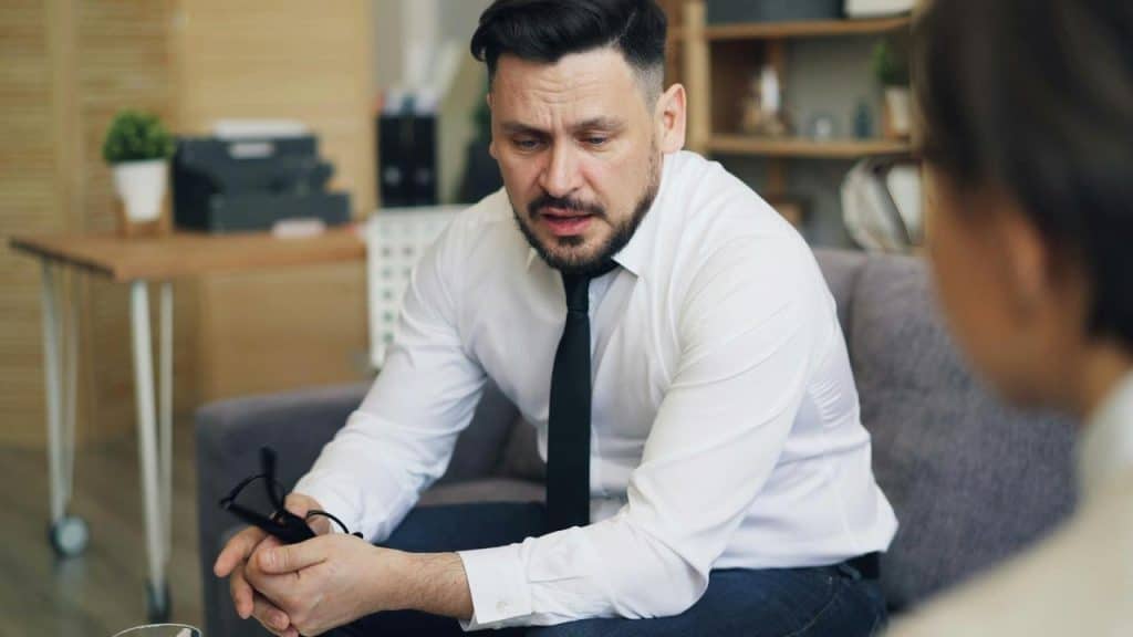 A man sitting on a couch holding his glasses while talking to someone.
