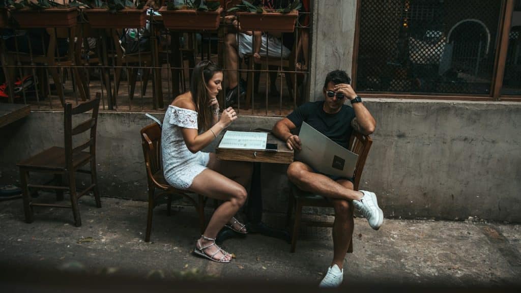 A man and a woman sitting at a small outdoor table, looking at menus.