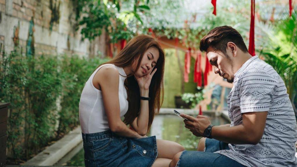 A woman looking upset while a man beside her looks at his phone.