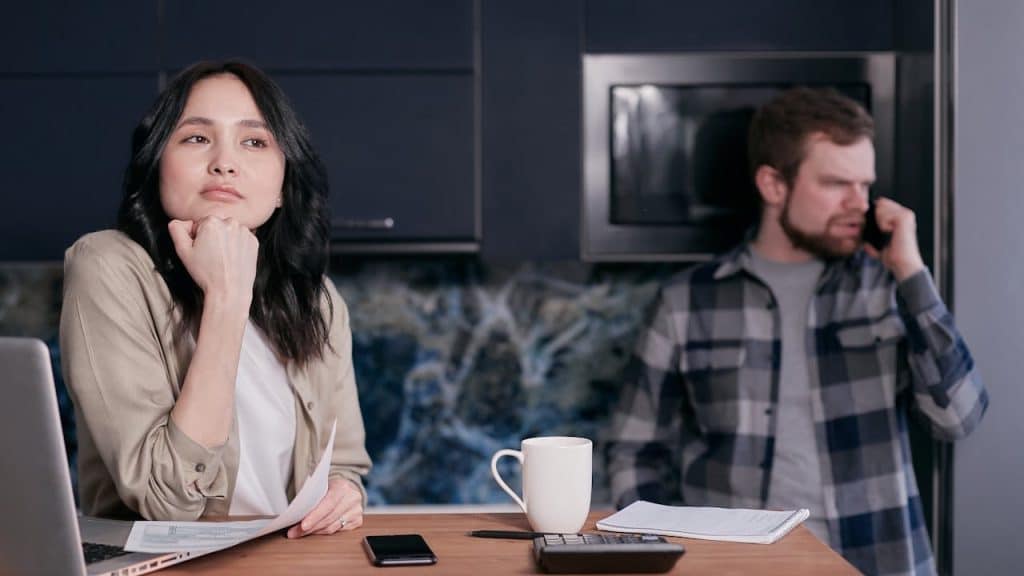 A Woman pondering over documents and a laptop in a kitchen, while a man talks on the phone in the background.