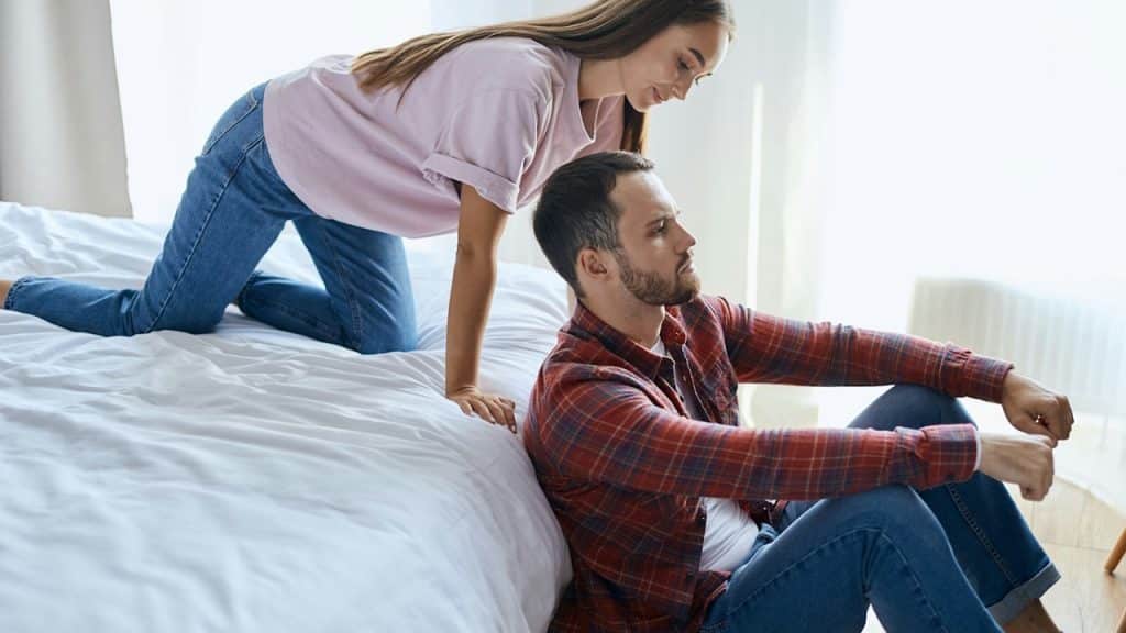 Woman kneeling on bed looking at man sitting on floor.
