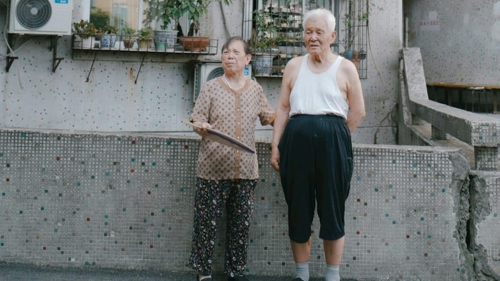 An old couple standing beside a gray concrete wall during daytime.