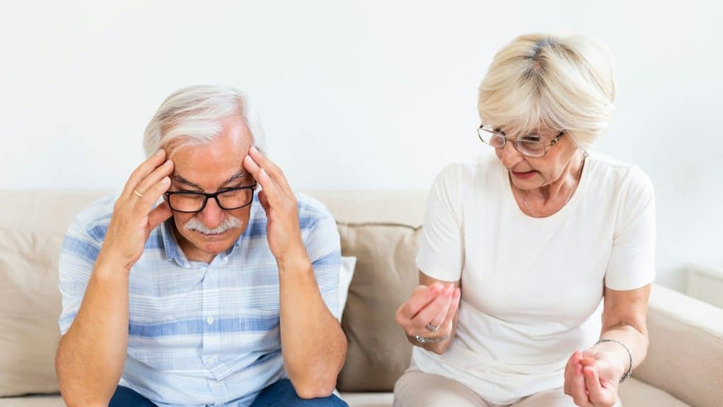 An elderly couple arguingβa senior man having a headache, holding his head between his hands.