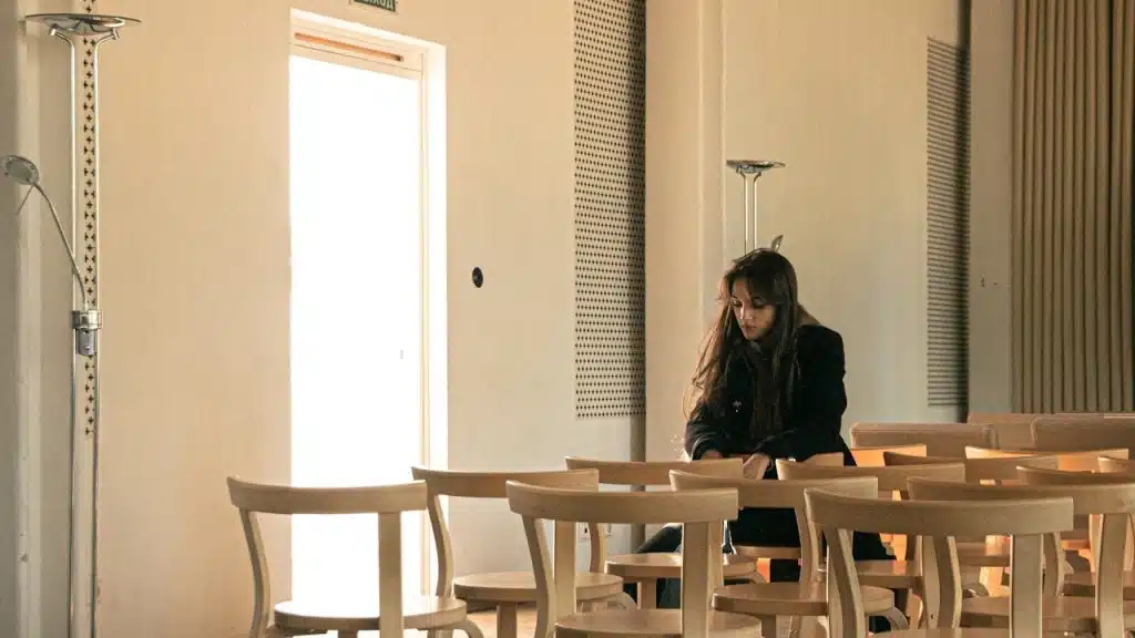 A woman sitting alone, looking downcast, in a brightly lit room filled with rows of empty wooden chairs.