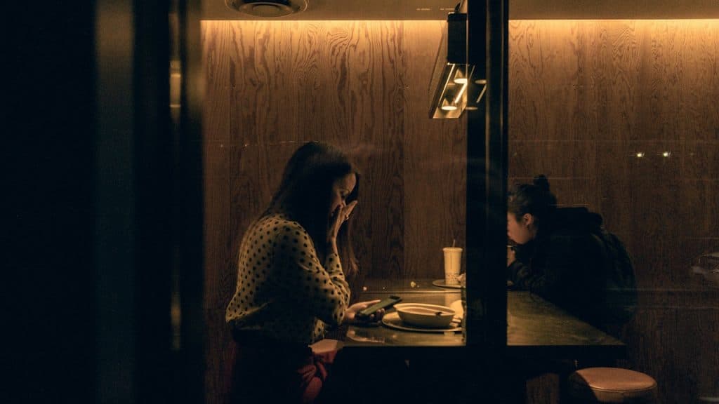 Two women sitting at a bar or table in a dimly lit, wood-paneled space, with one woman looking at her phone and the other eating.
