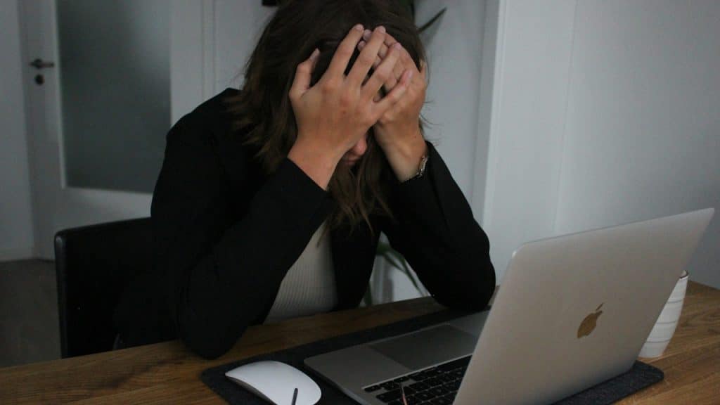Woman in a black blazer and white top, sitting at a wooden desk with a laptop, covering her face with both hands in frustration.