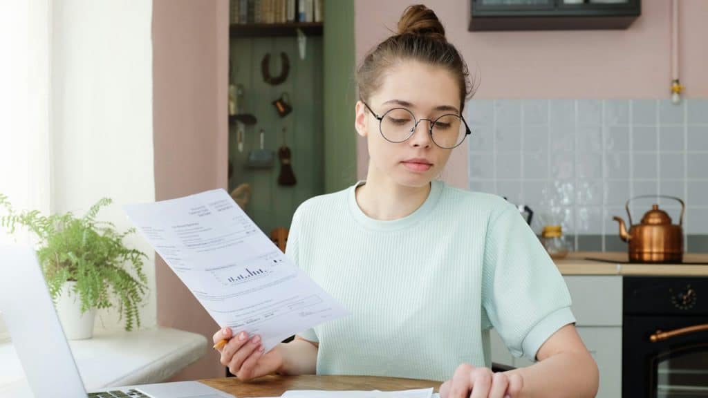 Young woman wearing round glasses and a light green shirt, sitting at a table with a laptop, looking at a document and using a calculator.