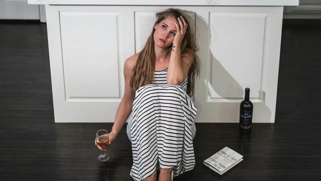 Woman in a striped dress sitting on the floor leaning against a white kitchen island, holding a glass of wine, with a bottle of wine and a paper nearby.