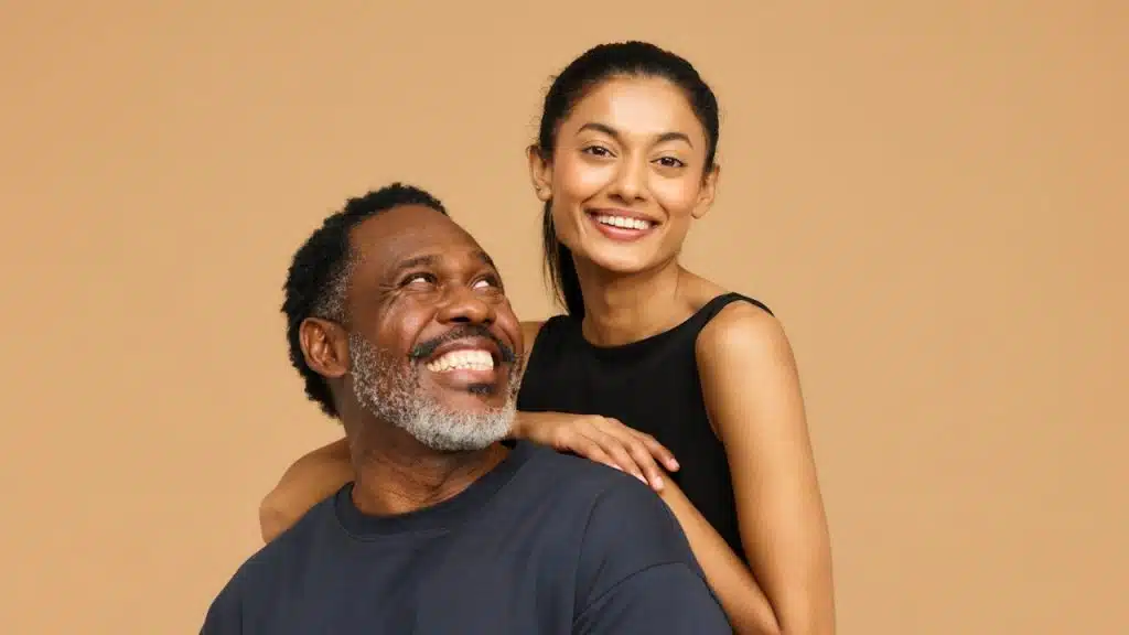 Smiling young woman in a black top standing behind an older Black man who is also smiling and looking upwards, against a plain tan background.