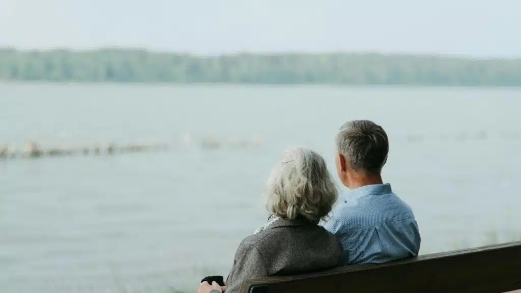 An older couple sitting on a bench and gazing out over a calm lake.