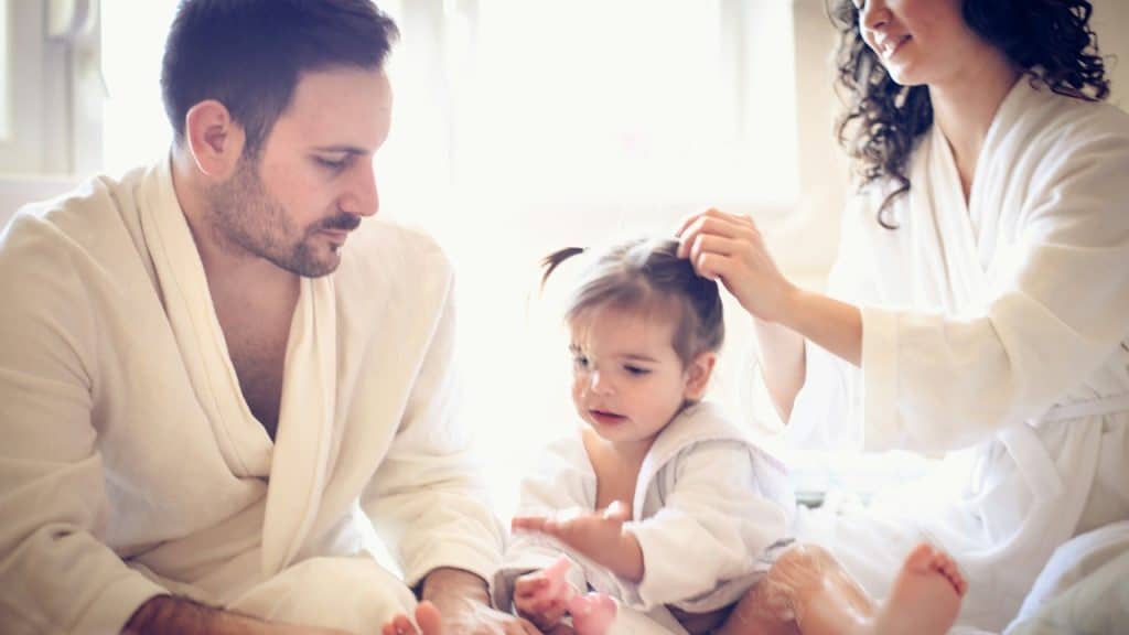 Parents in white robes sitting on a bed, with the father holding the feet of their young child, and the mother styling the child's hair.