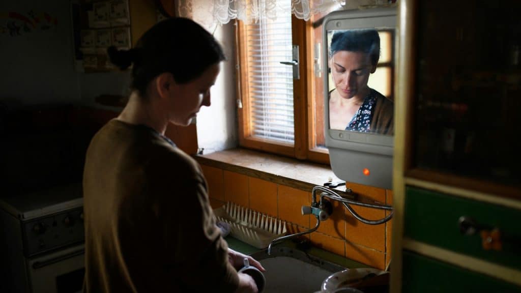Woman doing dishes in a dimly lit kitchen, with her reflection visible in an old television screen mounted nearby.