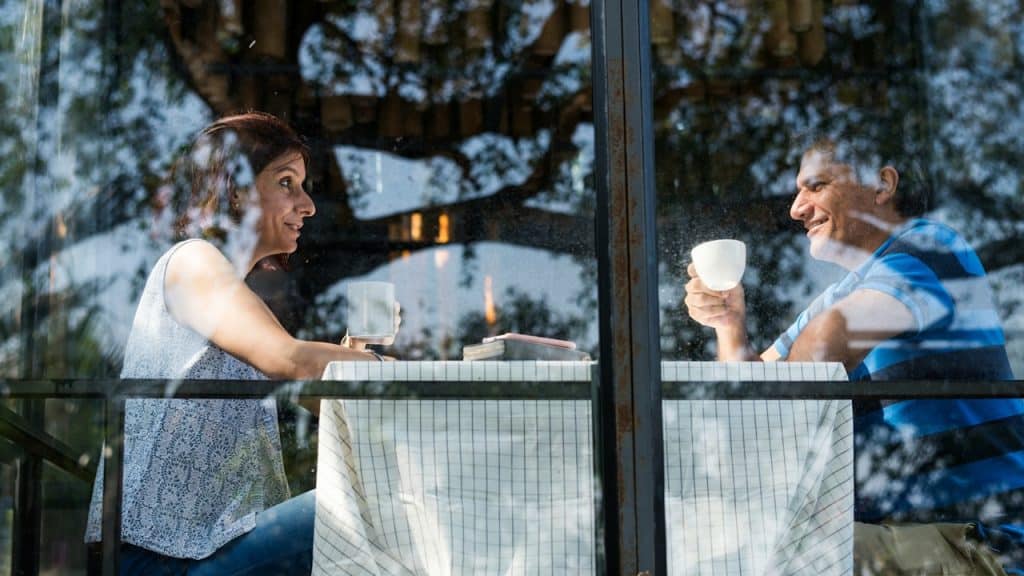 Middle-aged couple sitting at a small table behind a window, drinking coffee and smiling at each other, with trees reflected in the glass.
