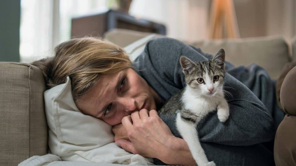 Woman lying on a couch with a small tabby and white kitten resting on her arm, looking towards the camera.