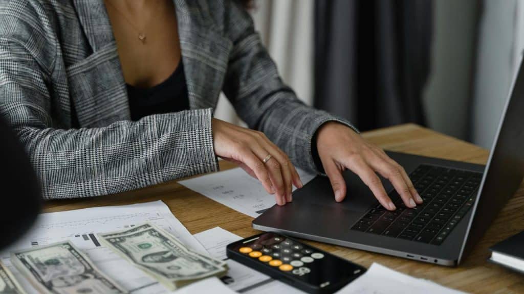 A woman working on a laptop with money, papers, and a calculator on the desk.