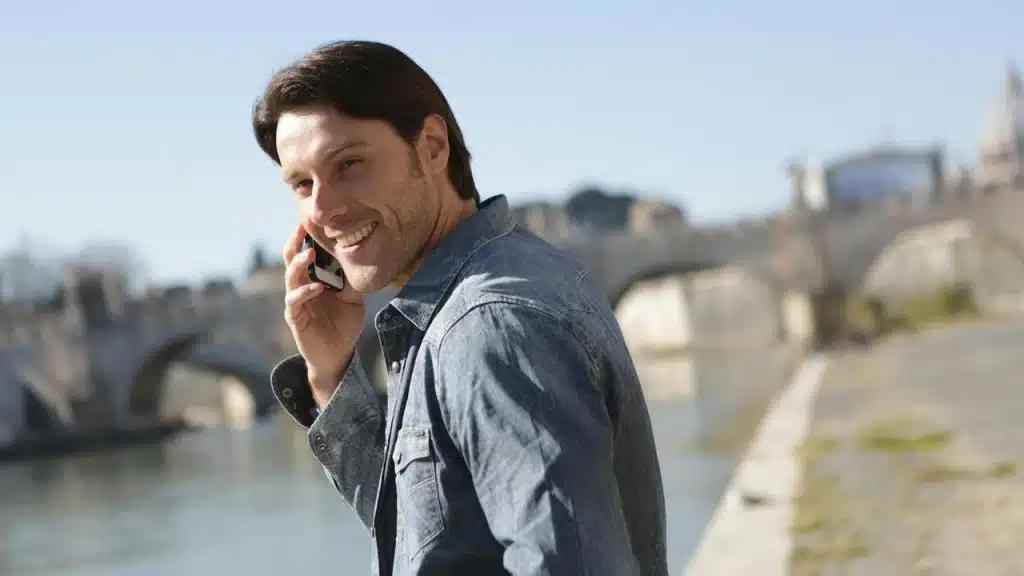 A smiling man talking on a phone while walking by a riverside.