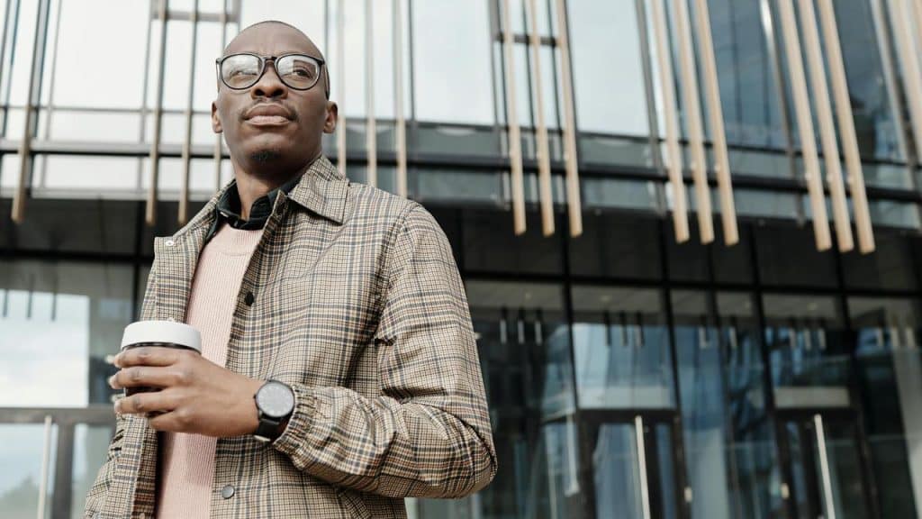 A man wearing glasses and a plaid jacket holding a coffee cup outside a modern building.