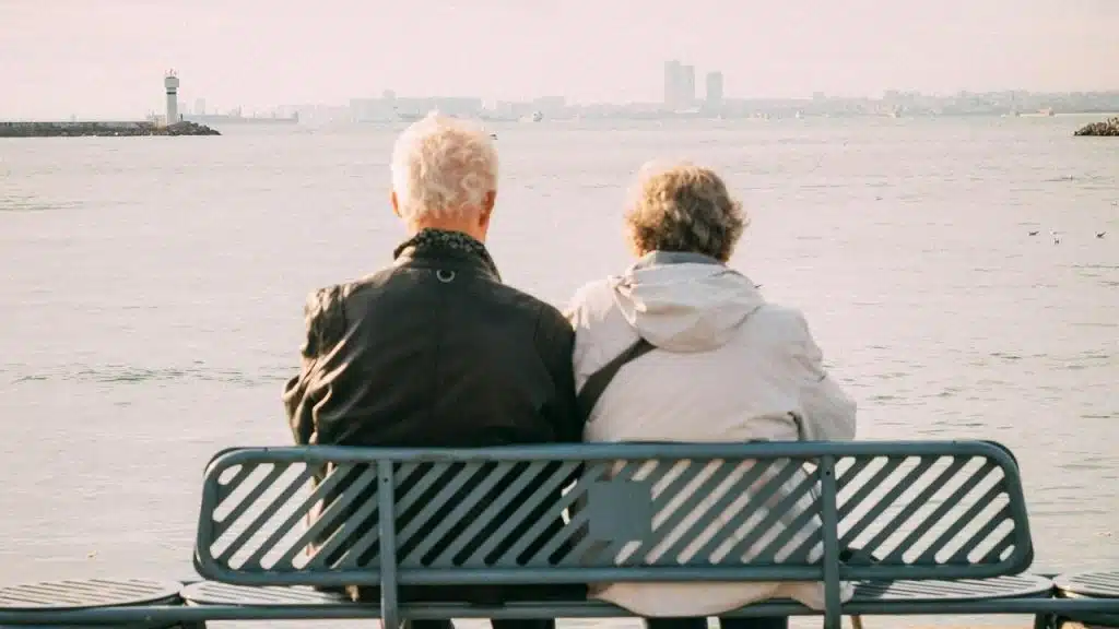 A senior couple sitting on a waterfront bench facing the sea.