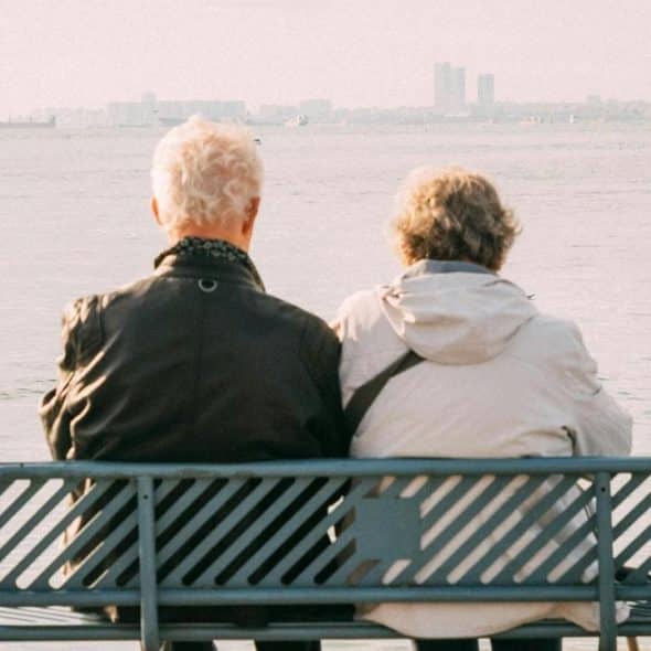 A senior couple sitting on a waterfront bench facing the sea.
