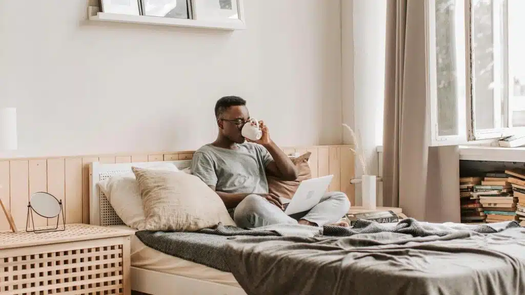 A man sitting on a bed drinking from a mug while using a laptop.