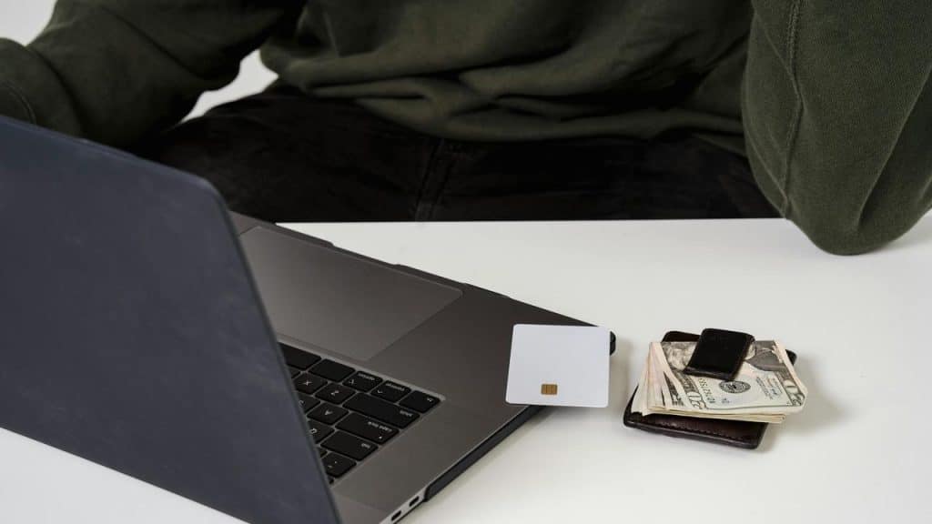 A person sitting at a desk with a laptop, a credit card, cash, and a wallet.